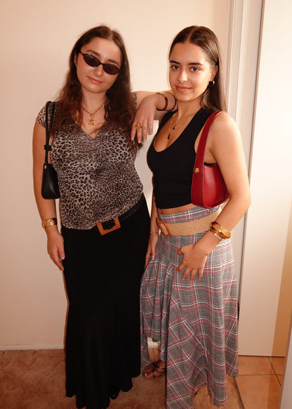Two women standing together indoors, one wearing sunglasses and a leopard print top, the other in a black top and plaid skirt.