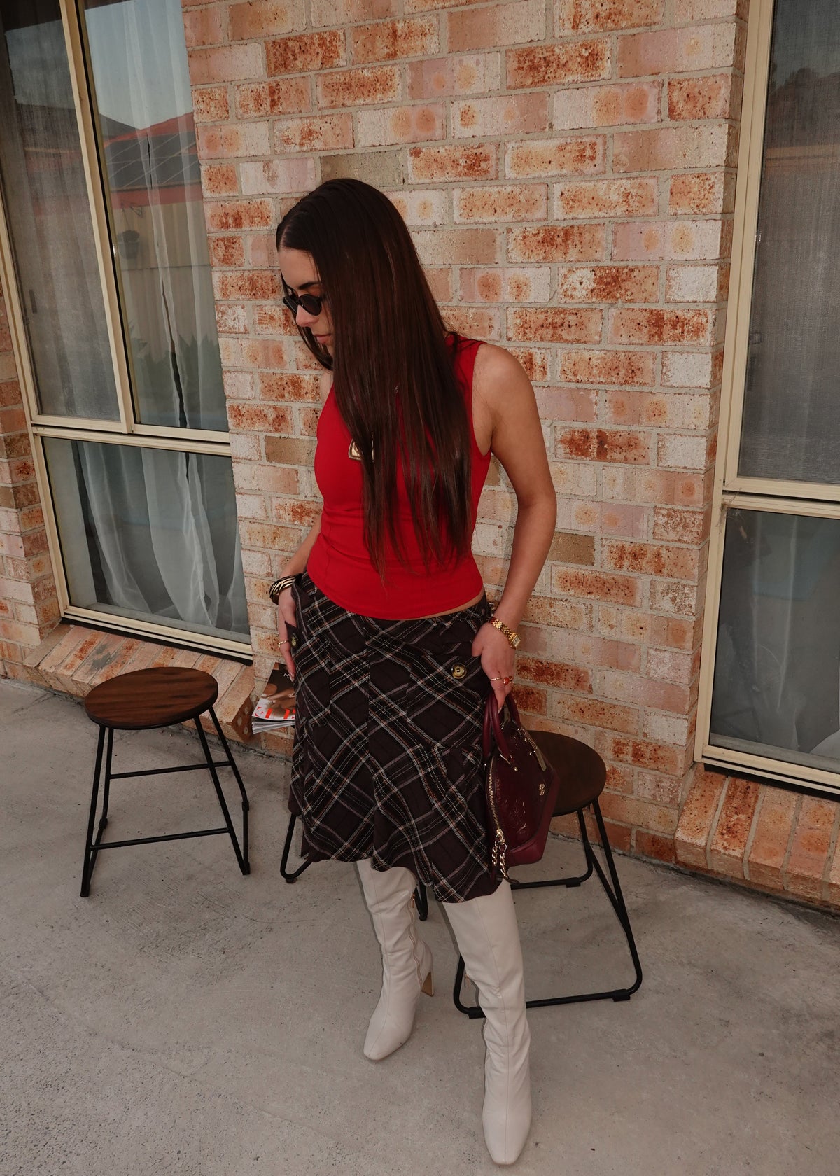 A woman wearing a red sleeveless top, a black and white plaid skirt, and white knee-high boots stands on a concrete patio in front of a brick wall.