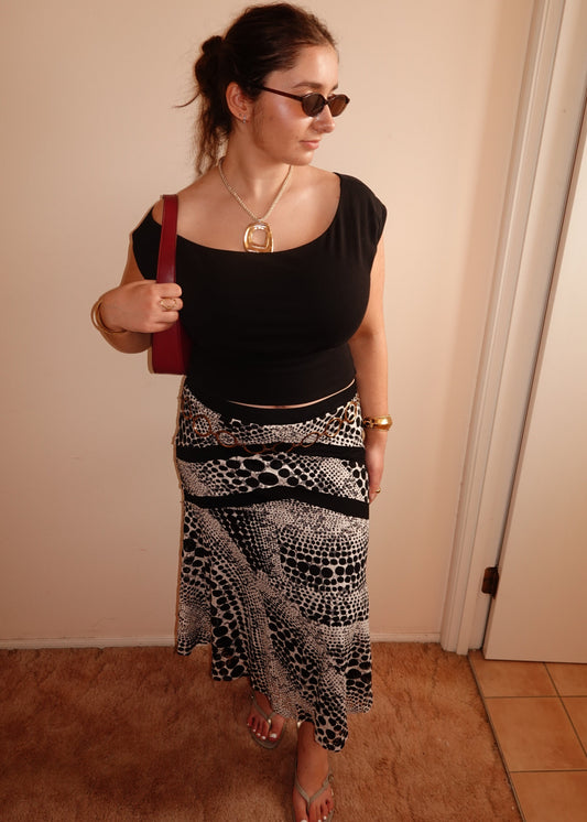 A woman wearing a black scoop-neck crop top and a black and white patterned midi skirt, accessorised with gold jewellery and sunglasses, standing indoors against a beige wall.