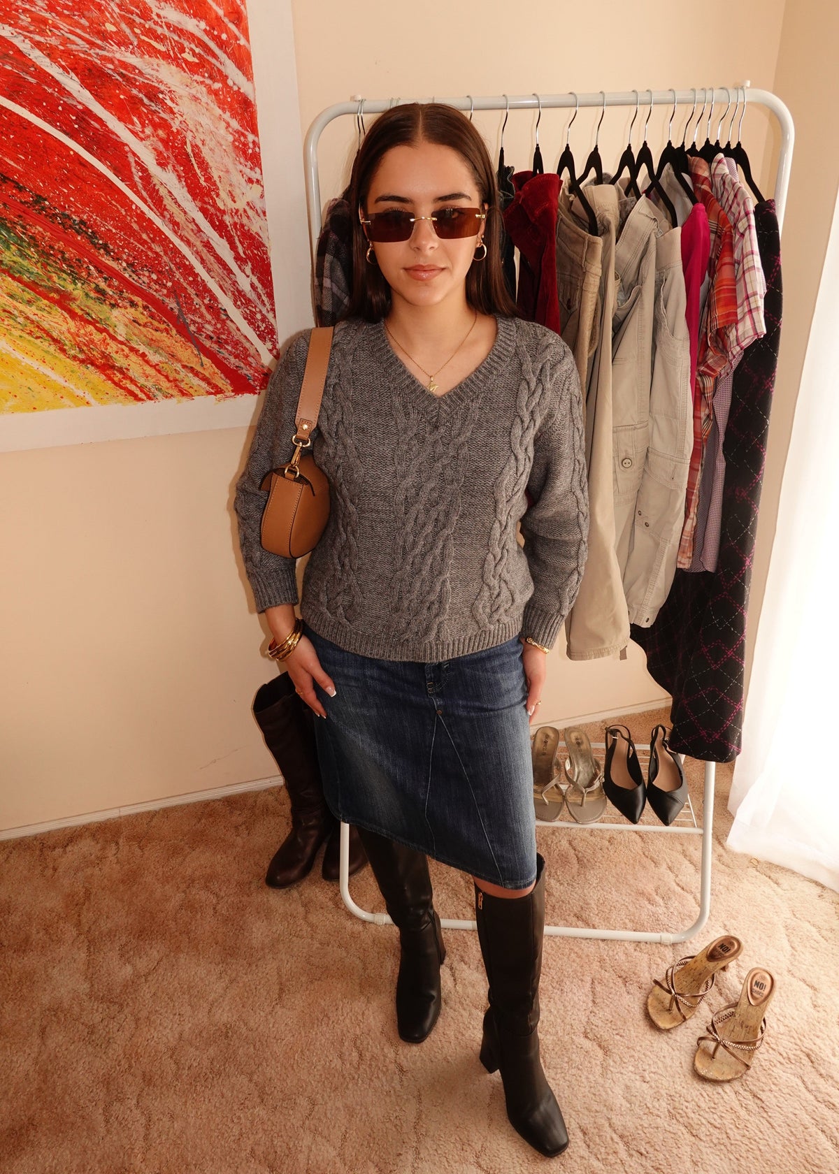 A woman wearing a gray cable-knit sweater, a blue denim skirt, and black knee-high boots stands in front of a clothing rack in a room with beige walls and a colorful abstract painting.