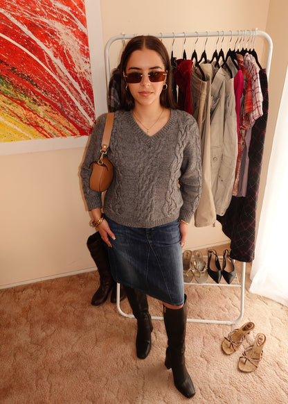 A woman wearing a gray cable-knit sweater, a blue denim skirt, and black knee-high boots stands in front of a clothing rack in a room with beige walls and a colorful abstract painting.